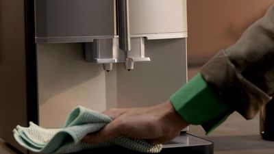 Une main avec un manchon vert essuie la base avant d'une machine à café automatique Siemens avec un chiffon doux.