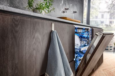 Lähikuva avoimesta, integroidusta astianpesukoneesta, jossa on sininen valo, modernissa keittiössä, jossa on tummat puupinnat.