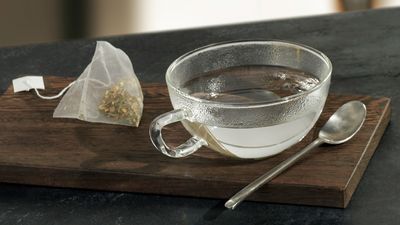 Tasse en verre contenant de l'eau chaude avec un sachet de th&eacute; et une cuill&egrave;re dispos&eacute;s &agrave; c&ocirc;t&eacute; sur un plateau en bois.