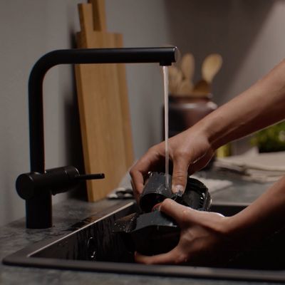 Personne rinçant les composants d'une machine à café sous un robinet noir moderne dans un évier de cuisine.