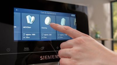 Un doigt effleurant l'écran tactile d'une machine à café Siemens, sélectionnant le programme de nettoyage calc'nClean dans la section Entretien & Maintenance du menu.