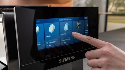 Un doigt effleurant l'écran tactile d'une machine à café Siemens, sélectionnant le programme de détartrage calc'nClean dans la section Entretien & Maintenance du menu.