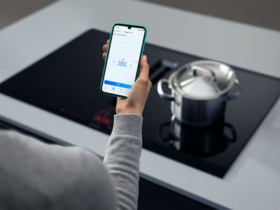 A person holds a smartphone above a stove, possibly checking a recipe or cooking instructions.