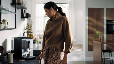 Une femme vêtue d'un pull marron se tient dans une cuisine moderne avec une machine expresso Siemens automatique sur le comptoir.