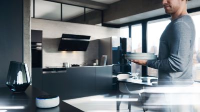 Un uomo in una cucina moderna tiene dei piatti, con un dispositivo per assistente vocale sul piano.