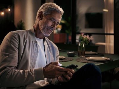 Ein Mann mit silbernem Haar sitzt an einem Esstisch und schaut auf sein Mobiltelefon.