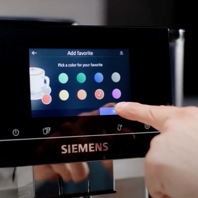 Un doigt effleurant l'écran tactile d'une machine à café Siemens, dans la section « Ajouter un favori » du menu sur l'écran.