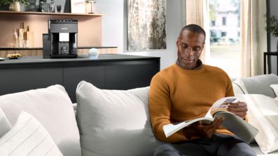 Homme lisant un magazine sur un canapé dans un salon moderne avec une machine à café Siemens sur un comptoir en arrière-plan.