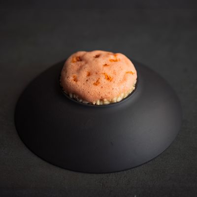 Un bol noir présente un plat crémeux surmonté d'une mousse mousseuse aux teintes orange et de notes de zeste d'orange sur une surface sombre.