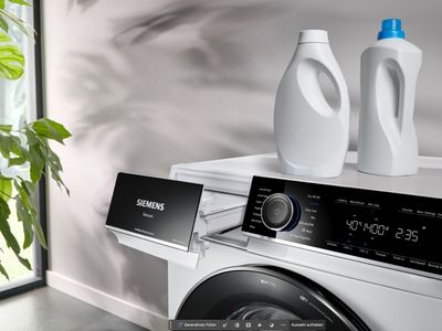 Deux bouteilles de lessive et d'assouplissant sont pos&eacute;es sur un lave-linge dont le tiroir est ouvert. &Agrave; c&ocirc;t&eacute; de la machine, une plante d'int&eacute;rieur verte projette l'ombre de ses feuilles sur le mur.