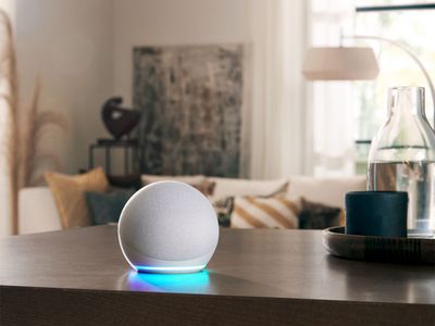 
       En grå Amazon Alexa smart høyttaler på et trebord med en sofa i stuen i bakgrunnen.
      