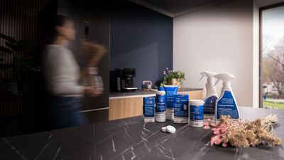 Uma mesa de cozinha com produtos de limpeza em destaque, uma pessoa em movimento e uma janela iluminada ao fundo. 