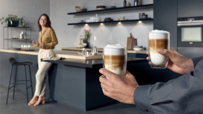 Au premier plan, deux mains apportent deux tasses de café latte à une femme appuyée à un comptoir de cuisine moderne en arrière-plan.
