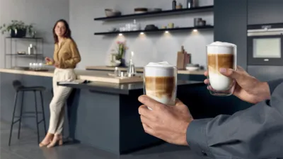 osoba trzyma w dloniach kawe latte , a tle kobieta oparta o blat kuchni