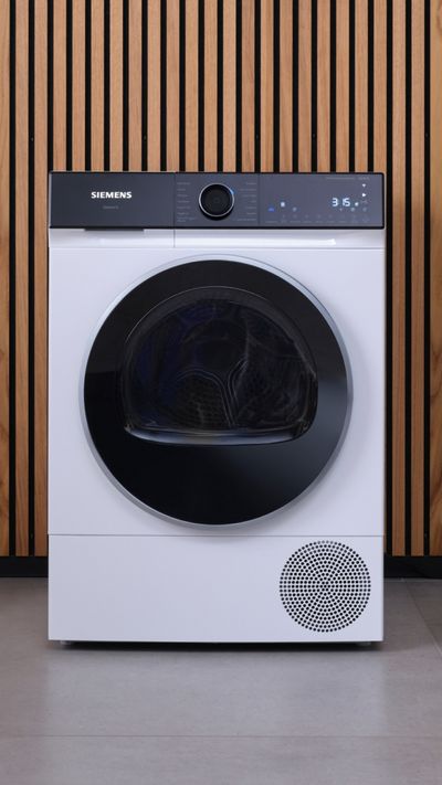 Ein Wärmepumpentrockner von Siemens steht vor einer holzverkleideten Wand.