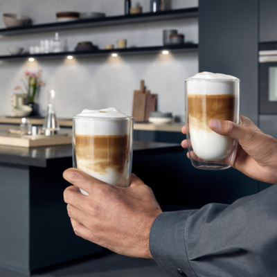 Deux mains tenant deux verres de latte macchiato devant un fond de cuisine.