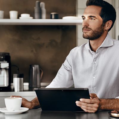 Un homme en chemise blanche assis &agrave; une table, tenant une tasse de caf&eacute; dans une main et une tablette intelligente dans l'autre.