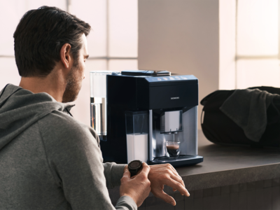 Een man in sportkleding staat voor een Siemens koffiemachine terwijl hij op zijn smartwatch kijkt.