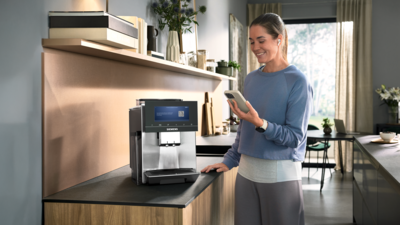 Femme utilisant l'application Siemens Home Électroménager sur un smartphone à côté d'une machine expresso automatique Siemens dans une cuisine élégante.
