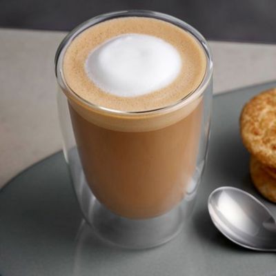 Un flat white dans une tasse en verre sur une assiette avec des biscuits et une cuillère.