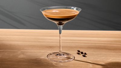Un verre &agrave; cocktail contenant un expresso martini est pos&eacute; sur une surface en bois clair &agrave; c&ocirc;t&eacute; de petits grains de caf&eacute; torr&eacute;fi&eacute;s.