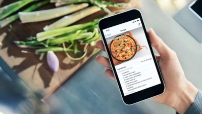 Detalle de un smartphone con una receta abierta en la app de Electrodom&eacute;sticos Siemens. Al fondo, una tabla de cortar con esp&aacute;rragos frescos 
