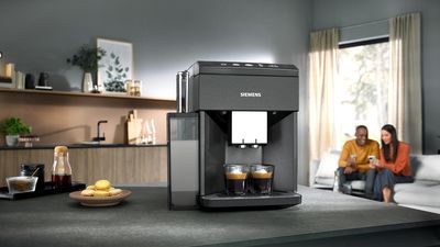 A Siemens coffee machine sits on a countertop, brewing two espresso shots, with macarons and beverages nearby, in a cozy living room setting.