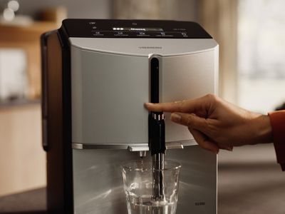 Osoba používající elegantní stříbrný kávovar k nalévání nápoje do průhledné sklenice s podsvícenými tlačítky nad ní.