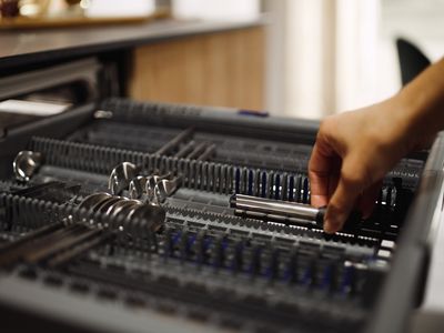 Ruka sáhne do zásuvky na nádobí a uspořádá nerezové příbory mezi úhledně uspořádaným náčiním.