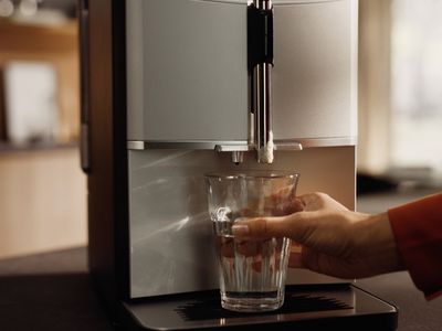 A hand holds a glass under a sleek silver water dispenser, preparing to fill it with water in a modern kitchen setting.