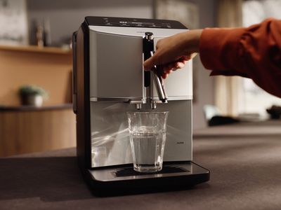 V útulném kuchyňském prostředí si člověk plní čirou sklenici vodou z elegantního moderního kávovaru.