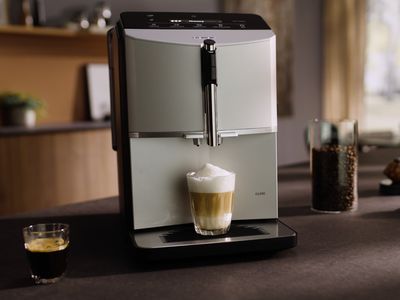 Elegantní kávovar v útulné kuchyni připravuje vrstvené latte s pěnou a zároveň tmavé espresso ve sklenici.