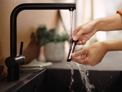 Osoba držící nástavec na kohoutek pod tekoucí vodou, s elegantním černým kuchyňským dřezem a jemnou zelení v pozadí.