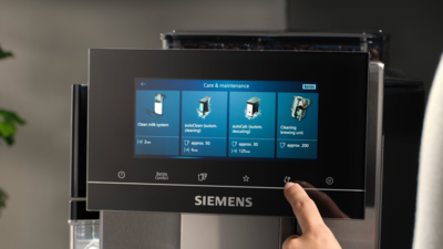 Un doigt effleurant l'écran tactile d'une machine à café Siemens, sélectionnant le programme autoCalc'n clean dans la section Entretien & Maintenance du menu.