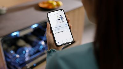 Eine Frau steht vor einem Siemens Geschirrspüler und blickt auf die smartStart Funktion auf einem Smartphone.
