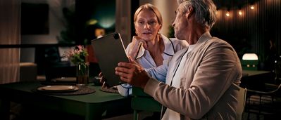 Una coppia di anziani seduta a un tavolo al chiuso, mentre guarda insieme un tablet in una stanza illuminata da luci calde e decorata con lucine a filo.