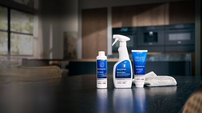 Tr&ecirc;s produtos de limpeza com r&oacute;tulos em azul sobre uma bancada em madeira com fundo escuro, acompanhado por um pano de limpeza branco, numa coxinha estilo moderno.