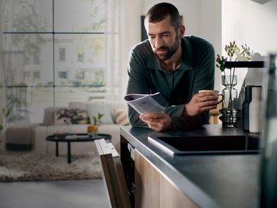 Man reading a brochure in a modern kitchen, leaning on a countertop with plants in the background.