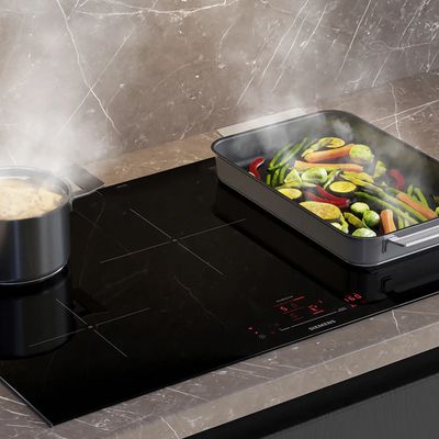 A pot of boiling pasta and a tray of vegetables cooking on a glossy ceramic induction cooktop.