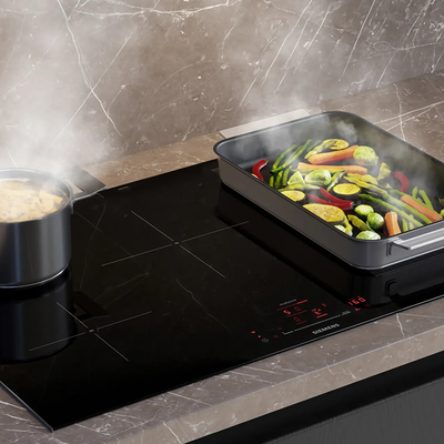Een pan met kokende pasta en een braadslede met groenten op een glanzende inductiekookplaat.