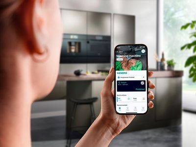 Uma pessoa segura um smartphone exibindo uma aplicação de máquina de lavar roupa numa cozinha moderna com eletrodomésticos elegantes e luz natural.