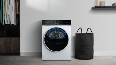 Uma máquina de secar Siemens com porta preta com tecnologia ReversibleDrumPlus posicionada ao lado de um armário cheio de roupa.