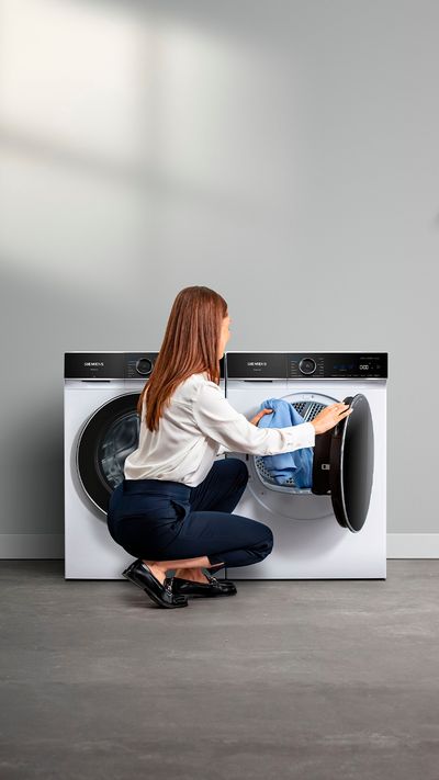 Uma mulher com visual profissional ajoelha-se para colocar roupa na máquina de lavar, com roupas penduradas nas proximidades e uma decoração moderna num espaço de lavandaria luminoso e minimalista.