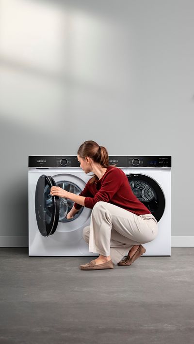 Žena v vínovém svetru se sklání, aby vložila prádlo do elegantní bílé pračky v moderní prádelně.