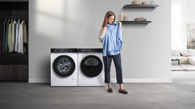 Žena drží modrou košili před moderní pračkou a sušičkou v minimalistické prádelně.