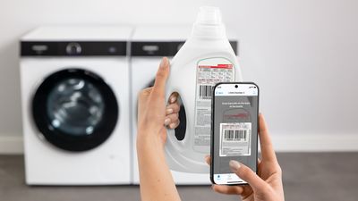 Osoba skenuje čárový kód lahve s pracím prostředkem chytrým telefonem, v pozadí pračka.