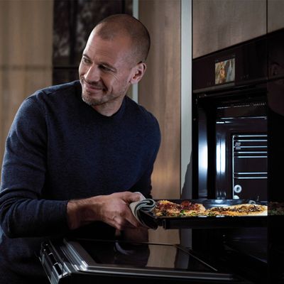 A person in a navy sweater pulls a baking tray with pizza from a modern oven in a stylish kitchen.