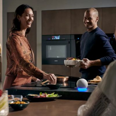A man and woman stand in a modern kitchen, with a smart speaker labeled 'Alexa' visible on the counter.  