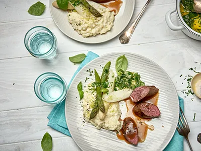 Cremiges Risotto, zartes Fleisch mit Sauce und Spargel, serviert mit Wasser auf einem rustikalen Tisch.
