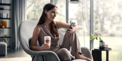 A woman in a casual outfit relaxes in an armchair, holding a coffee cup and a smartphone, with natural light streaming in through large windows.